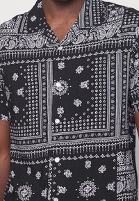 Homme portant une chemise noire à manches courtes boutonnée avec un imprimé blanc paisley et bandana géométrique.