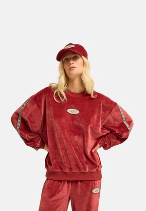 Blonde woman wearing a red corduroy sweatshirt and pants set with patterned stripes on sleeves and a matching red cap.