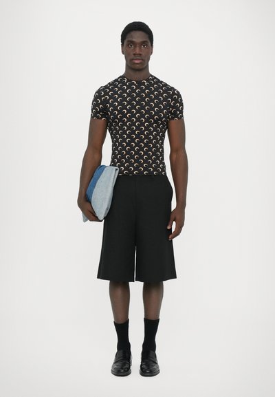 Black shirt with a repeating crescent pattern, paired with knee-length black shorts. Holding a folded light blue denim item, wearing black socks and shoes.