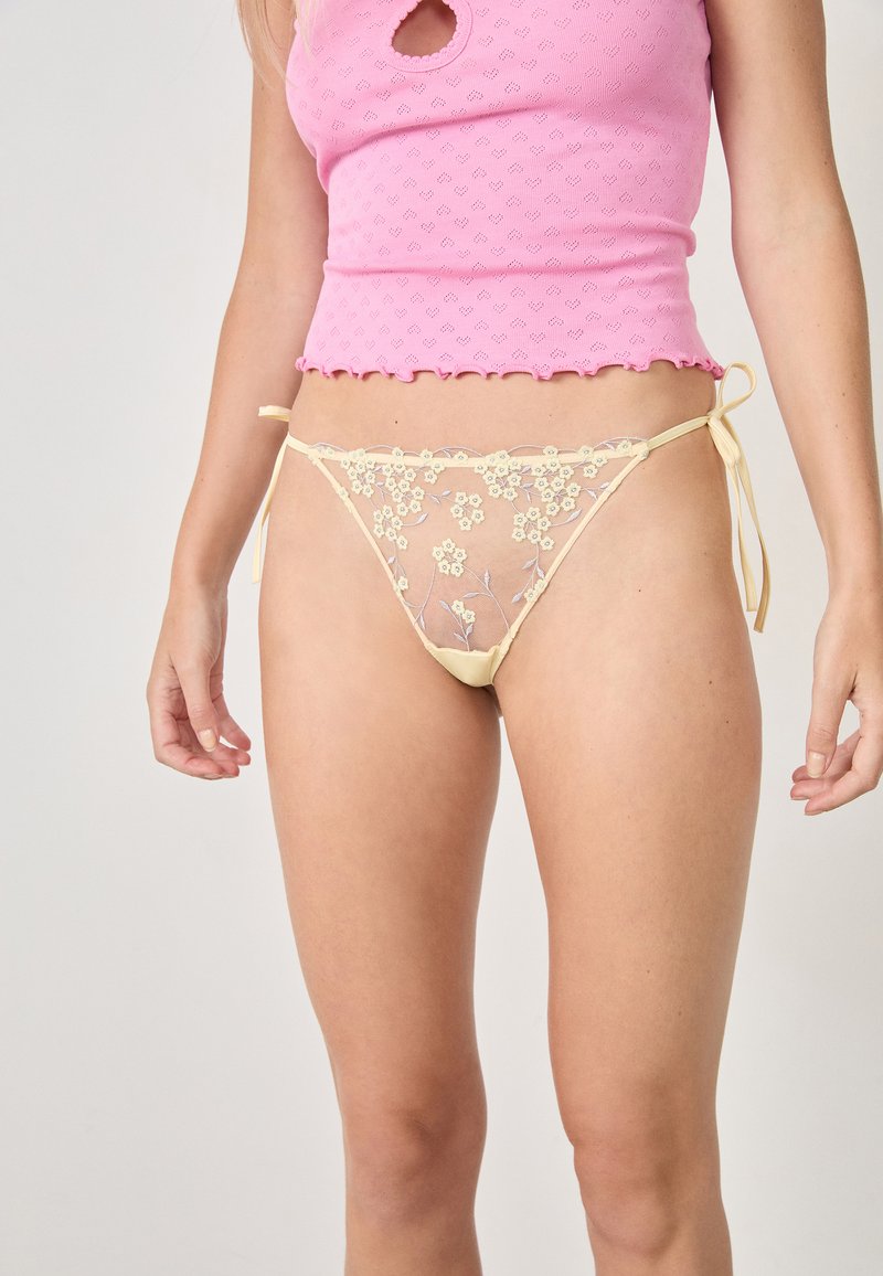 Tanga de encaje amarillo semitransparente con bordado floral, con lazos laterales y un corte de cintura baja. Acompañado de un top corto texturizado en color rosa.