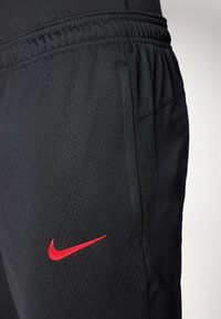 Crne Nike trenirke s elastičnom gumom u struku i bočnim džepovima. Crveni Nike swoosh logo na lijevom bedru. Glatka, lagana tekstura materijala.