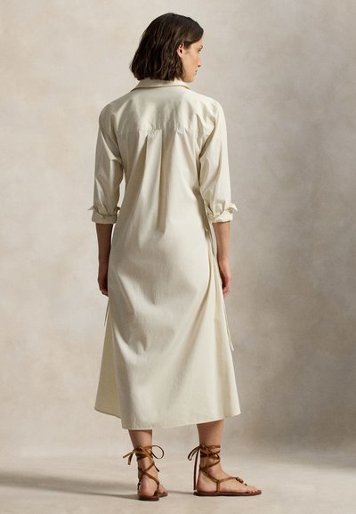 Φόρεμα μακρυμάνικο σε ανοιχτό μπεζ χρώμα με γιακά και μανίκια που είναι σηκωμένα, με πλαϊνές σχισμές και ρέουσα σιλουέτα. Καφέ σανδάλια με λουριά.