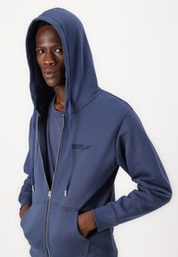 Marineblå zip-hoodie med en lomme foran, snøre i hætten og broderet "REPLAY" logo. Blødt stof, afslappet pasform, sømløst design.