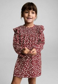 Punainen kukallinen mekko valkoisilla yksityiskohdilla, pitkät hihat ja rypytetyt olkapäät. Mukana on rypytetty vartalo-osa ja löysähelmainen hame.