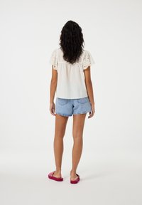 Witte blouse met pofmouwen, met perforaties, gecombineerd met high-waisted denim shorts. Voeten in roze fluffy sandalen. Neutrale achtergrond.
