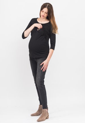 Schwarzes Mutterschaftsoberteil mit drapiertem Design, das einen verdeckten Stillzugang bietet. Kombiniert mit dunklen Jeans und braunen Chelsea-Stiefeln.