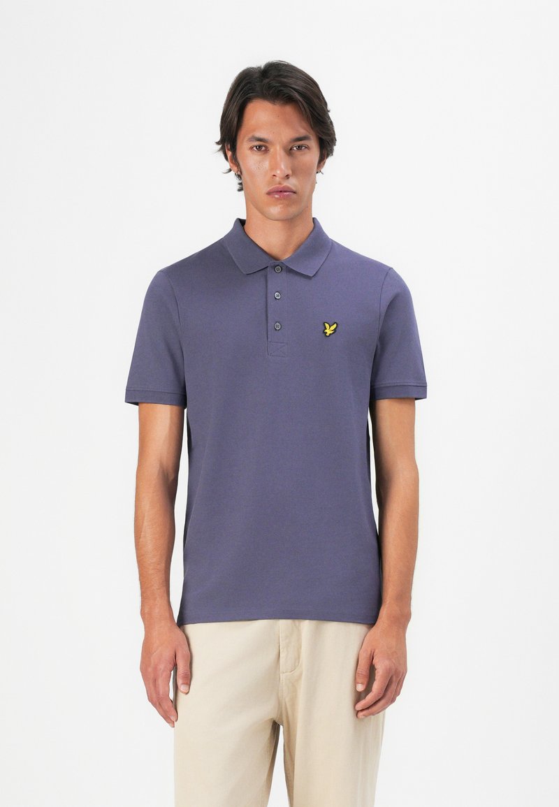 Lilla bomullspolo med kort erme, tre knapper foran, og en gul brodert logo på venstre bryst. Slim fit design.