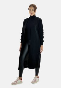 Cardigan lungo nero con un motivo di maglia testurizzata, indossato sopra un dolcevita nero, abbinato a leggings neri lucidi e sneaker chiare con dettagli rosa.