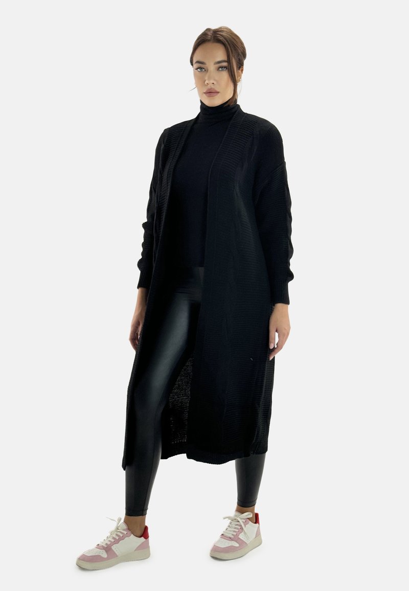 Cardigan lungo nero con un motivo di maglia testurizzata, indossato sopra un dolcevita nero, abbinato a leggings neri lucidi e sneaker chiare con dettagli rosa.