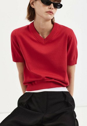 Person trägt schwarze Sonnenbrille, einen roten kurzärmligen V-Ausschnitt Pullover über einem weißen Hemd und schwarze Hosen, sitzt vor einem einfarbigen hellen Hintergrund.