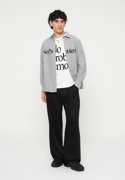 Graues Hemd über einem weißen T-Shirt mit schwarzem Text, kombiniert mit schwarzen weit geschnittenen Hosen und schwarzen Sneakers.