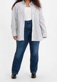 Kvinna iklädd lösa svarta stövlar, blå jeans, vit linne och en överstor vit skjorta med svarta vertikala ränder mot en enfärgad bakgrund.