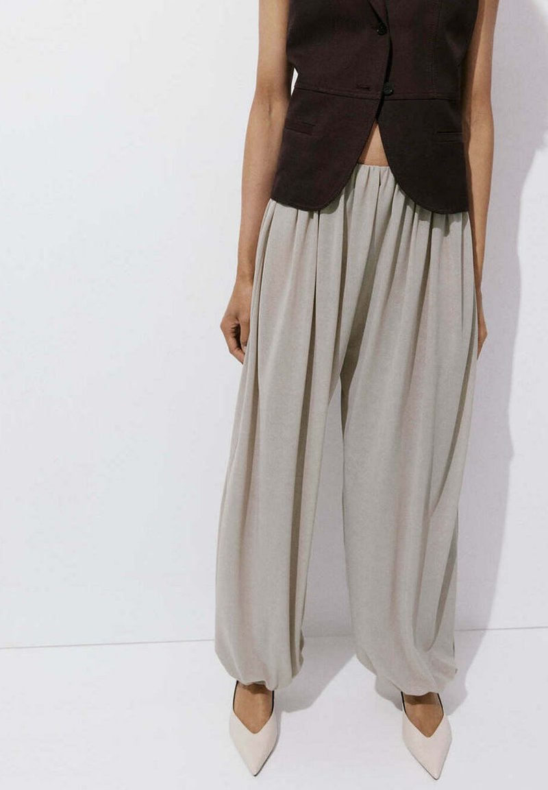 Mannequin portant un pantalon large ample beige clair avec des ourlets froncés, associé à un gilet noir ajusté sans manches et des talons blancs pointus.