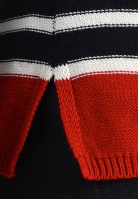 Bawełniany sweter w kolorze granatowym z wzorami w białe i czerwone paski. Designerski charakter podkreślają ściągacz u dołu oraz boczne rozporki. Widoczna jest pleciona faktura dzianiny.