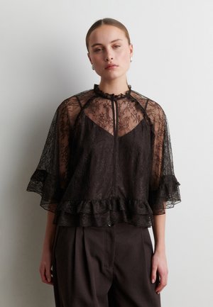 Jeune femme aux cheveux tirés en arrière porte un chemisier noir en dentelle transparente avec des manches et un ourlet volantés, associé à un pantalon sombre sur fond blanc.
