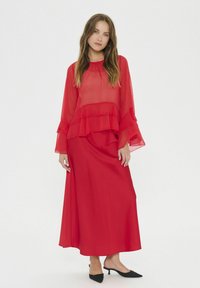 Rotes Maxikleid mit geschichteten, transparenten Rüschenärmeln und glatter Textur, kombiniert mit schwarzen spitzen High Heels.