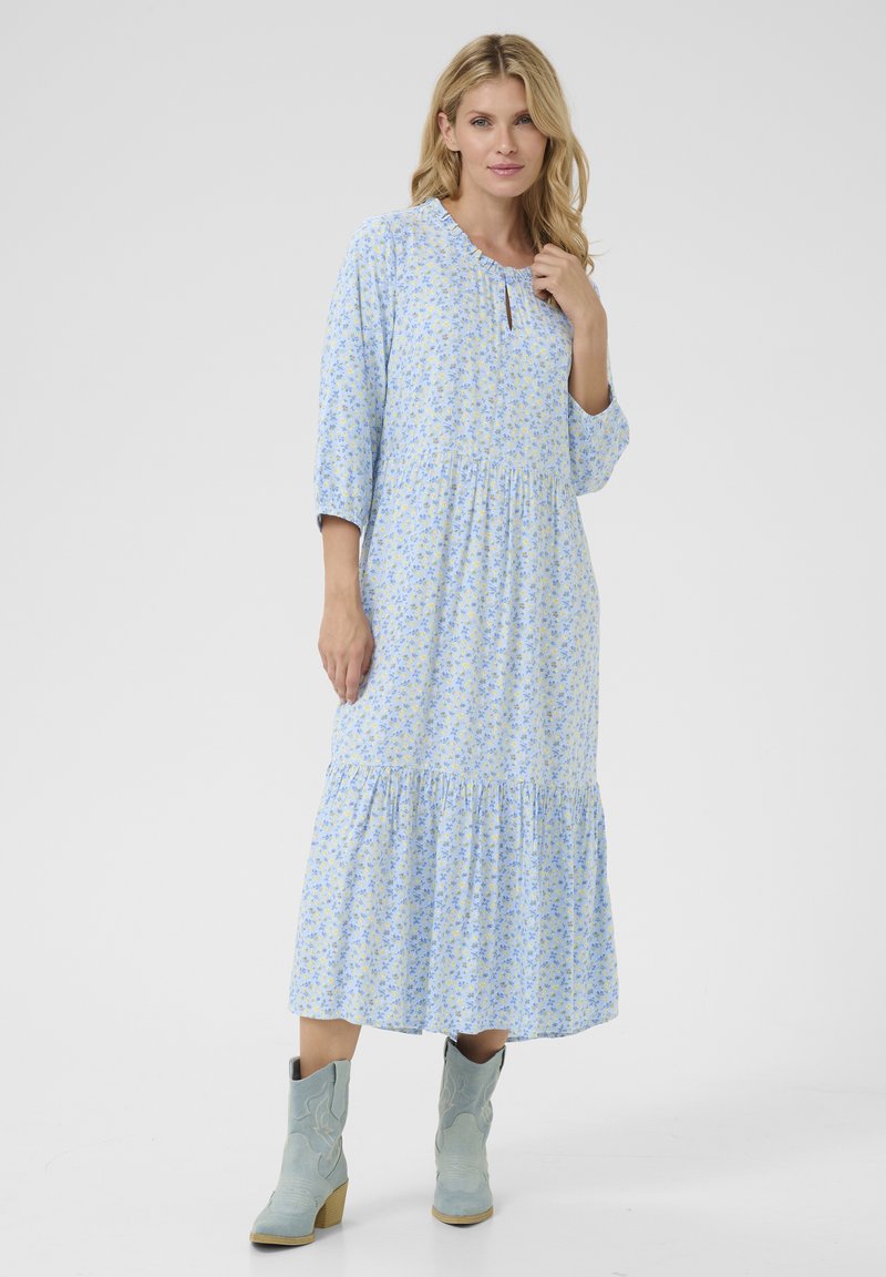 Zila ziedu maksi kleita ar trim ceturtdaļa piedurknēm, viļņotu apakšmalu un atvērtu kakla detaļu. Valkāta ar gaiši ziliem kovboju zābakiem.