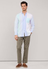 Lichtblauw en wit gestreept overhemd met een patchzak; gecombineerd met beige broek en bruine instappers, staand tegen een neutrale achtergrond.