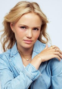 Guldsmycken med delikata kedjor, pärlor och en fjärilsdetalj, i kombination med en ljus denimjacka som framhäver detaljer på armarna.