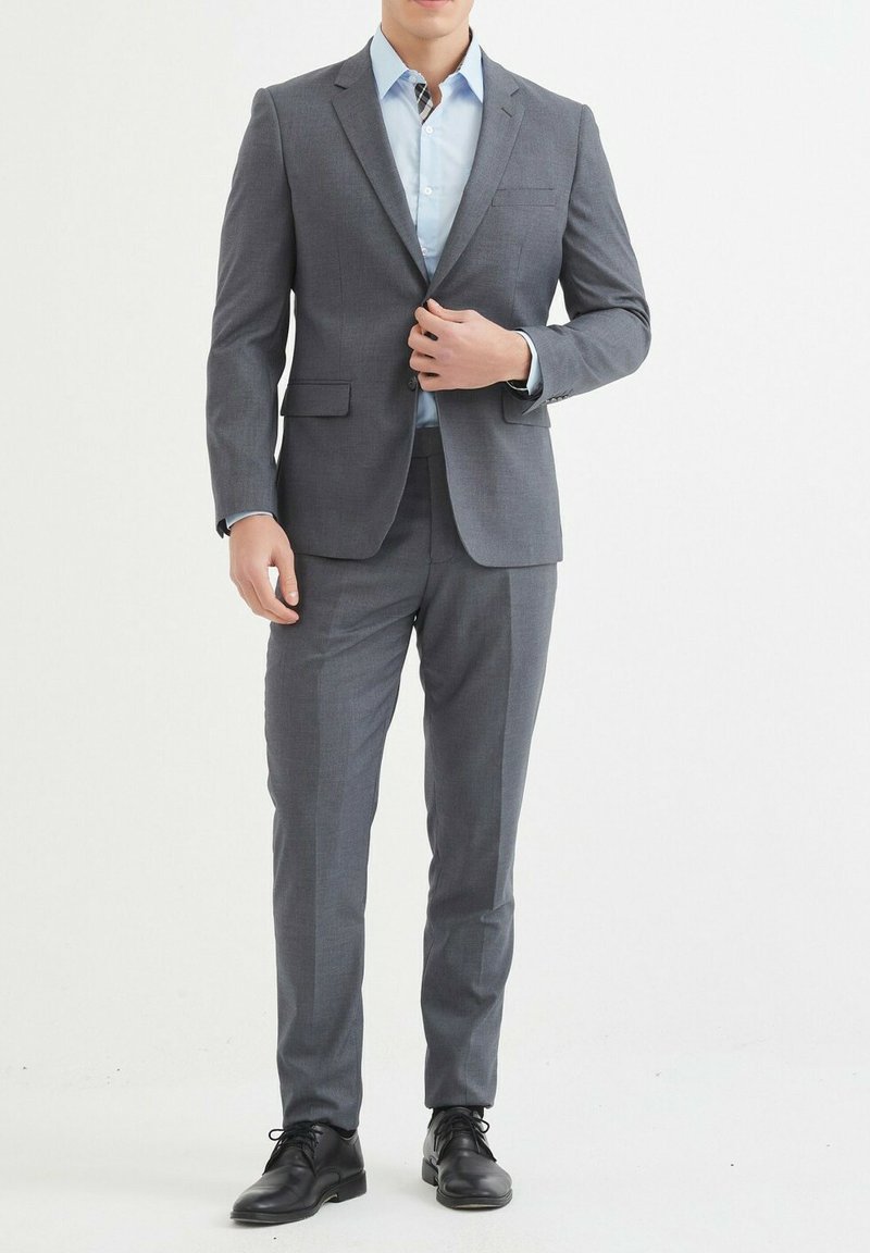 Costume gris à coupe ajustée, blazer à un bouton, pantalon assorti et chemise bleu clair. Des chaussures de ville noires complètent la tenue.