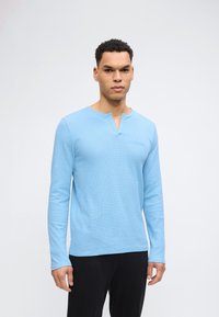 Άνδρας με κοντά κατσαρά μαλλιά, φορώντας ανοιχτό μπλε μακρυμάνικη μπλούζα με υφή βάφλας και μαύρο παντελόνι, μπροστά από ουδέτερο φόντο.