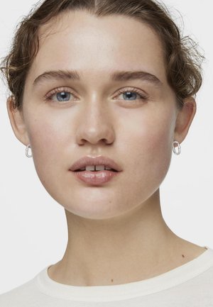 Aretes de aro plateados, textura suave, forma ovalada; modelo con piel clara y cabello rizado castaño claro, vistiendo una camisa blanca.