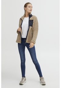 Veste en polaire beige avec des accents noirs, fermeture éclair, col montant et deux poches latérales. Associée à un jean bleu ajusté et des baskets claires.