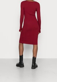 Robe en tricot bordeaux à manches longues et silhouette ajustée. Caractérisée par une texture côtelée et une longueur mi-mollet, portée avec bottines noires.