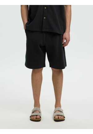 Personne portant un short noir, une chemise noire boutonnée et des sandales gris clair sur un fond uni.