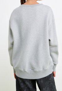 Felpa grigia con vestibilità morbida, polsini e orlo a costine, realizzata in morbido cotone. La vista posteriore mostra dettagli minimi e un colletto classico.