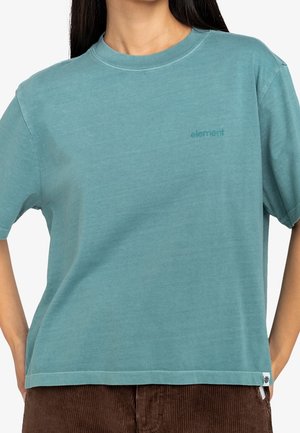 Femme portant un T-shirt à manches courtes bleu sarcelle avec le logo « element » sur la poitrine et un pantalon en velours côtelé marron, les mains posées sur les hanches.