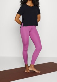 Top negro de manga corta, de corte relajado, combinado con leggings ajustados de color rosa vibrante. Diseño sencillo, textura suave, usado sobre una esterilla de yoga marrón.