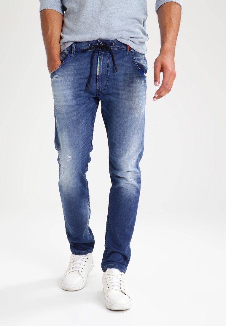 Hombre vestido con jeans azules desgastados con cintura ajustable, combinados con zapatillas blancas y una camiseta de manga larga gris claro, caminando hacia adelante.