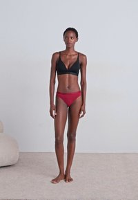 Zwarte driehoekige beha en rode hooggesneden bikini onderkant. Glad materiaal, minimalistisch ontwerp. Model staat op een licht textuur tapijt tegen een effen muur.