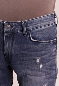 Vaqueros de mezclilla en azul oscuro con un acabado desgastado, que cuentan con cinco bolsillos, un cierre con botones y una textura ligeramente desvaída en los muslos.