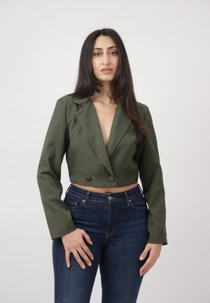 Frau steht vor einem einfarbigen Hintergrund, trägt ein kurzes olivgrünes Blazer und dunkelblaue Jeans, blickt nach vorne und hat die Hände entspannt.