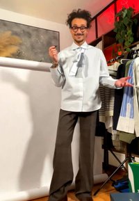 Muž s kudrnatými vlasy a brýlemi stojí u stojanu na oblečení, má na sobě bílou košili s velkou mašlí a hnědé široké kalhoty uvnitř.