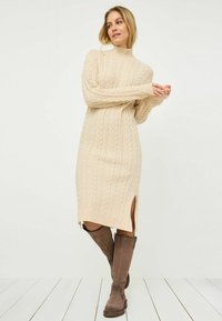 Vestito a maglia beige con maniche lunghe, lunghezza fino a metà coscia e spacchi laterali. Abbinato a stivali marroni alti fino al ginocchio.