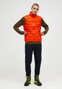 Gilet imbottito arancione con un motivo a onde trapuntato, colletto alto, chiusura a zip. Indossato sopra una maglietta marrone a maniche lunghe, abbinato a pantaloni neri e stivali verdi.