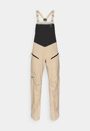 Beige wasserdichte Overalls mit einem schwarzen oberen Bereich, verstellbaren Trägern, seitlichen Reißverschlusstaschen und verstärkten Bündchen für Langlebigkeit.