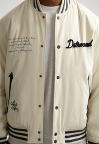 Lys beige bomberjakke med navyblå stribet ribkrave og manchetter. Har broderet tekst og motiver i sort. Lukning med trykknapper.