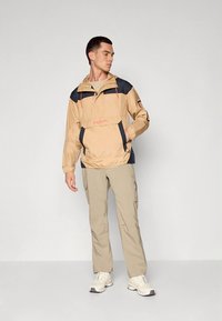 Beige windbreaker met marineblauwe accenten, voorkant met zak, verstelbare capuchon en elastische manchetten, gecombineerd met lichtgekleurde cargobroek en sneakers.