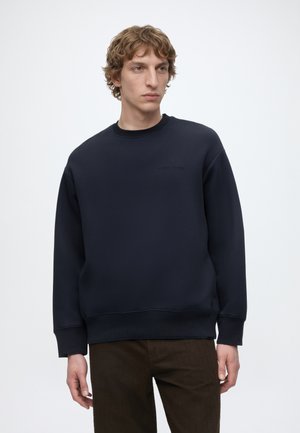Jeune homme aux cheveux bouclés portant un sweat-shirt bleu marine uni et un pantalon foncé, debout sur un fond clair uni.