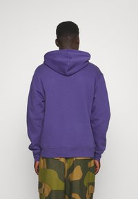 Sudadera morada con una gran capucha, puños y dobladillo acanalados. La tela parece suave y duradera. Combinada con pantalones cargo de estampado de camuflaje.