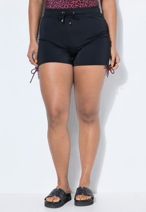 Vrouw draagt zwarte shorts met trekkoorden en zijstrikjes, staat blootsvoets in zwarte slippers met oranje gelakte teennagels, op een witte achtergrond.