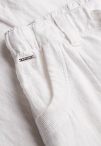 Pantalon en lin blanc avec taille élastique, poche latérale, passants de ceinture et petite étiquette métallique de la marque "Superdry" près de la poche.