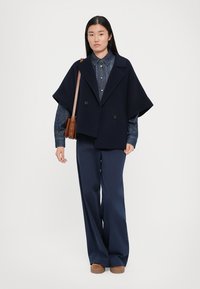 Marineblauwe wolmix cape met dubbele knoopsluiting, oversized pasvorm, gecombineerd met een spijkerhemd, wijd uitlopende marineblauwe broek en tan platform schoenen.