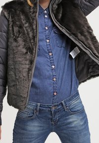 Kamizelka z sztucznego futra w ciemnoszarym kolorze, zapinana na zamek z przodu, noszona na niebieskiej koszuli z guzikami. Połączona z niebieskimi dżinsami z kontrastowym szwem.