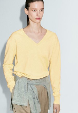 Maglione giallo a V con maniche lunghe, in tessuto lavorato a maglia. Wrap grigio attorno alla vita, abbinato a pantaloni beige.