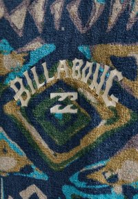 Tekstil med tekstur, der har et abstrakt mønster i blå, grøn og brun; inkluderer et cremegult broderet "BILLABONG" logo med et stiliseret tal "23."